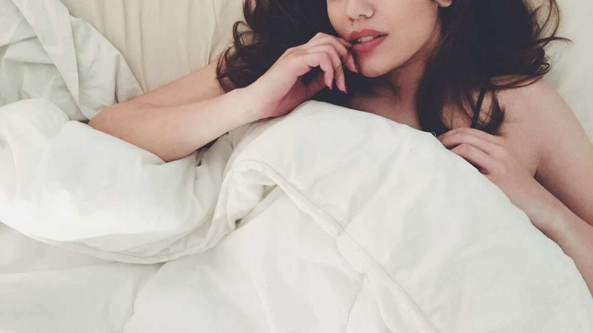 A female escort with long, dark hair, tucked into bed in her Los Angeles incall apartment.