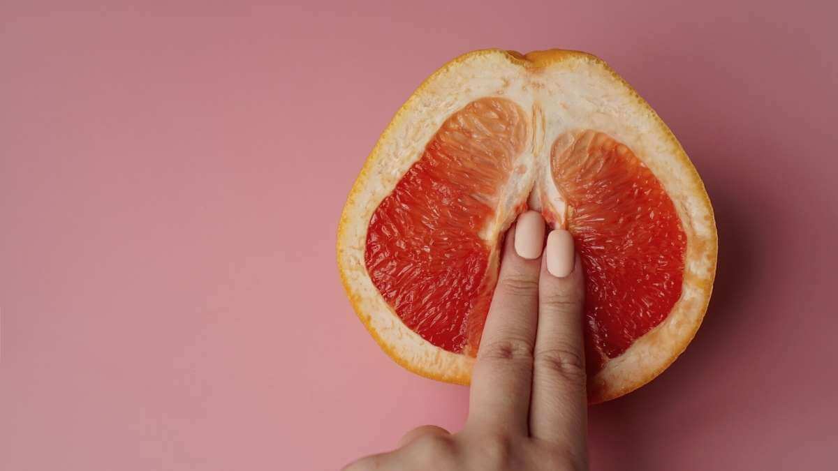 A female escort with pink nails touches half an orange on a pink background.