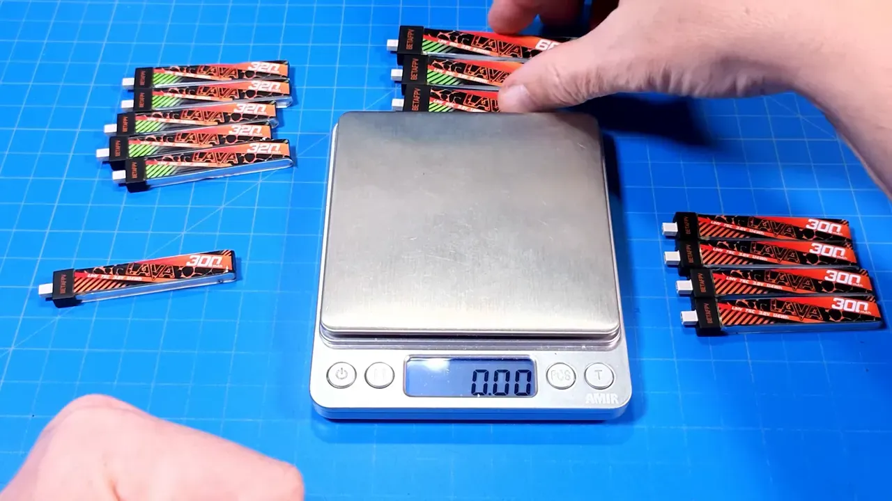 Top-down view of a digital scale on a blue grid mat with BetaFPV Lava II 320mAh (green label) and original Lava 300mAh packs arranged around it, hand placing packs