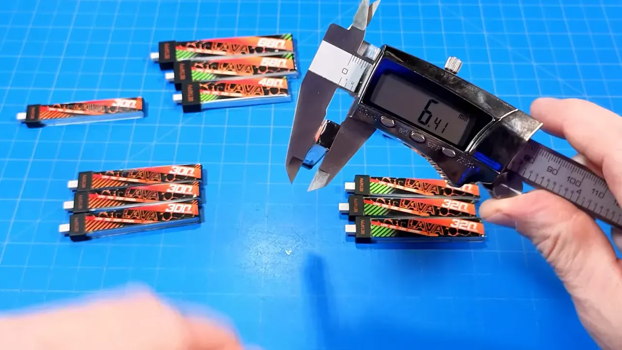 Digital calipers measuring a BetaFPV Lava battery showing a 6.41 mm readout, with stacks of Lava and Lava II packs arranged on a blue grid mat