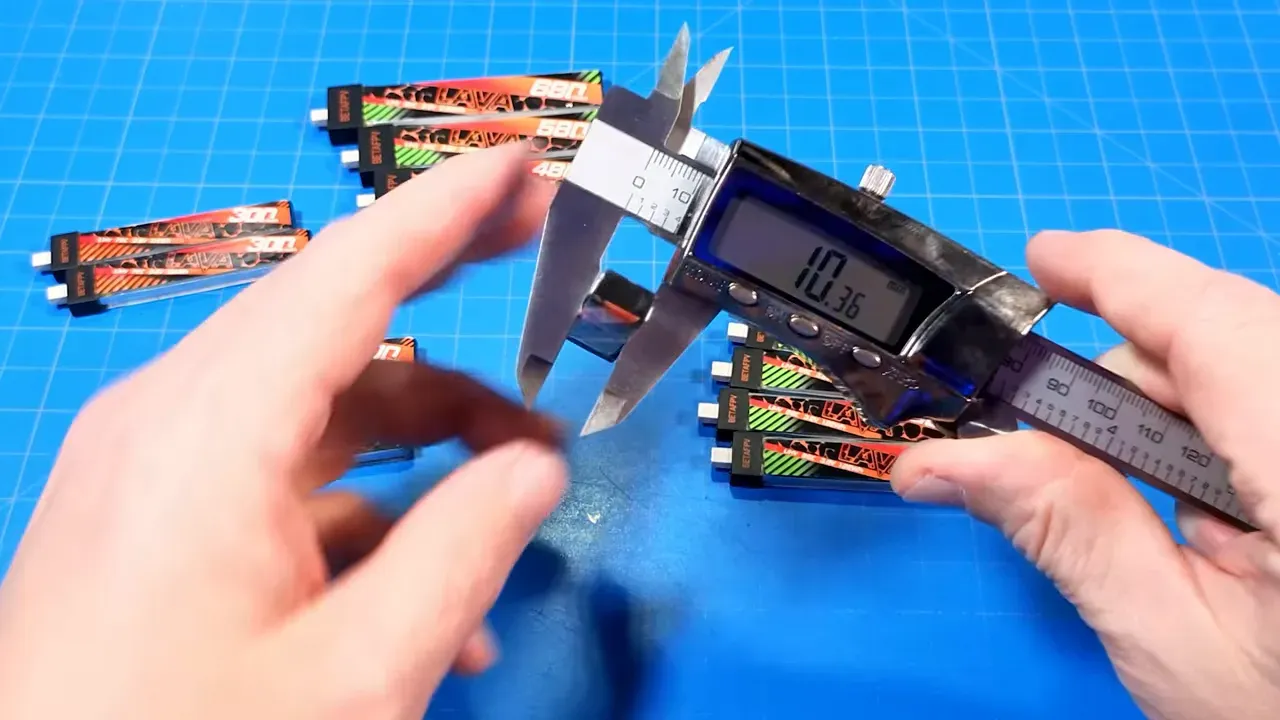 Close-up of digital calipers measuring a Lava battery pack on a blue grid mat with stacks of original and Lava II packs visible, display reading 10.36 mm
