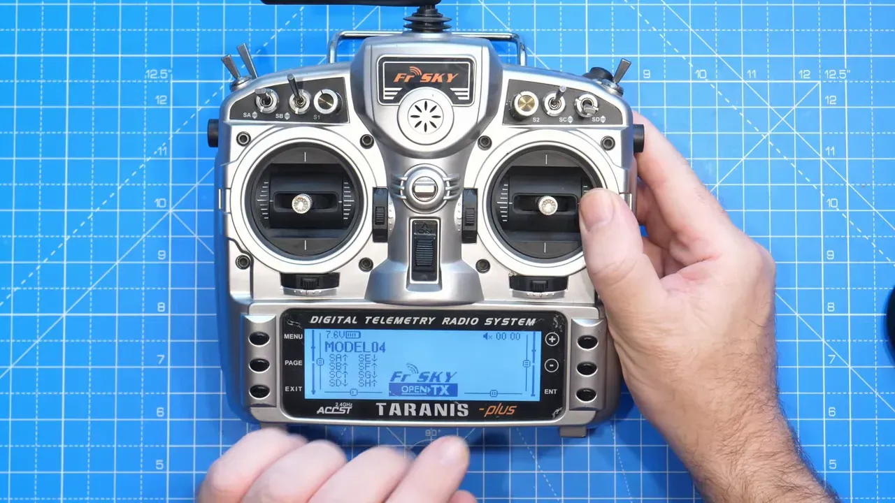 Top-down, high-clarity image of a FrSky Taranis transmitter on a blue cutting mat showing the monochrome model screen and both hands for scale