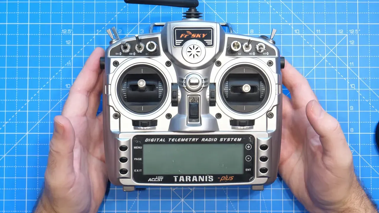 Top-down view of a FrSky Taranis transmitter centered on a blue grid mat, showing gimbals, switches and display clearly