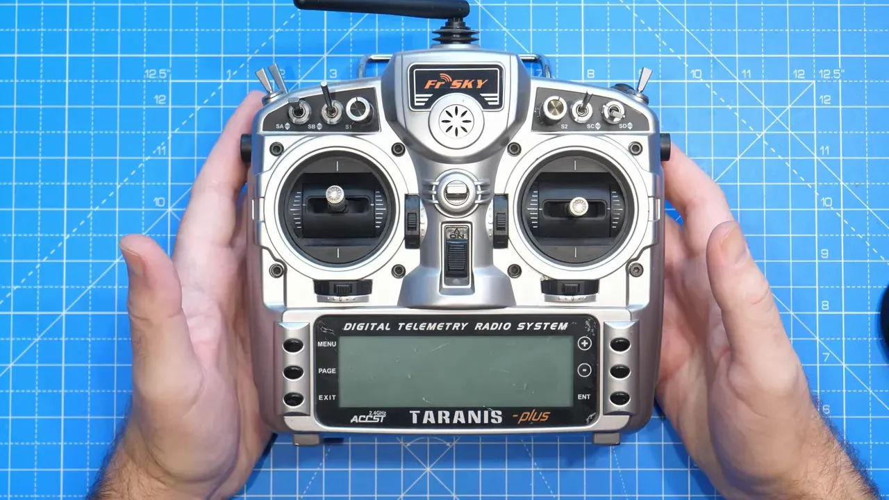 Top-down high-resolution view of a FrSky Taranis transmitter on a blue grid mat with hands visible, showing gimbals, switches, antenna and front display