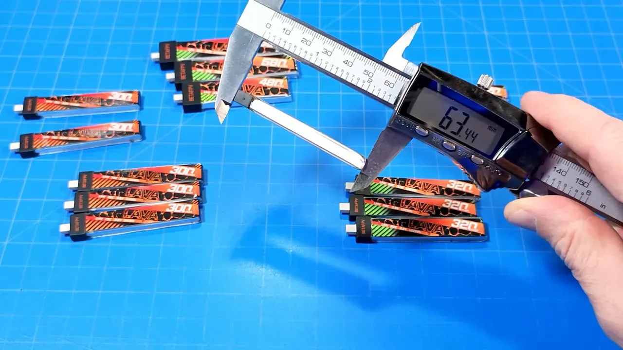 Digital calipers reading 63.44 mm while measuring the length of a Lava 320mAh battery on a blue grid mat with stacks of original and Lava II packs visible.