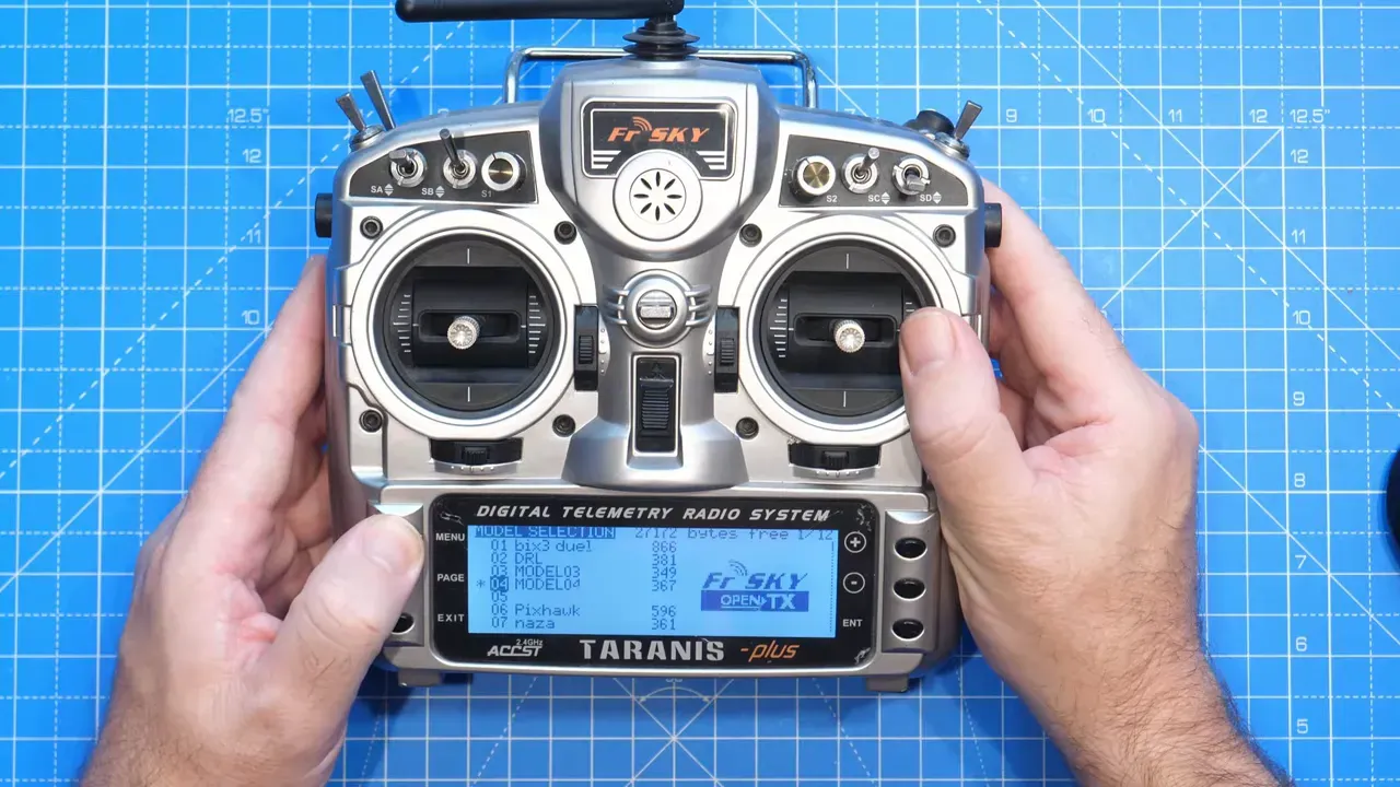 Top-down view of a FrSky Taranis transmitter with the model selection menu visible on the monochrome screen and hands framing the radio
