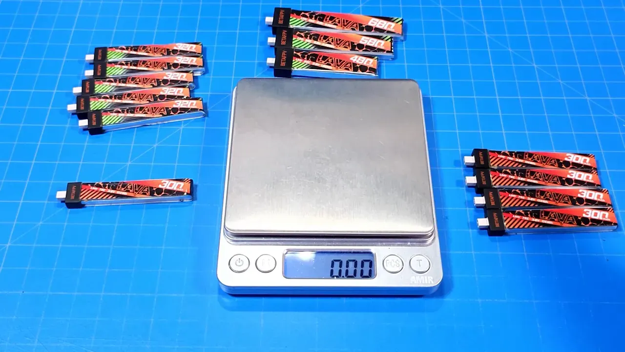 Top-down view of a silver digital scale on a blue grid mat with BetaFPV Lava II 320mAh (green label) and original Lava 300mAh packs arranged around it, scale display reading 0.00