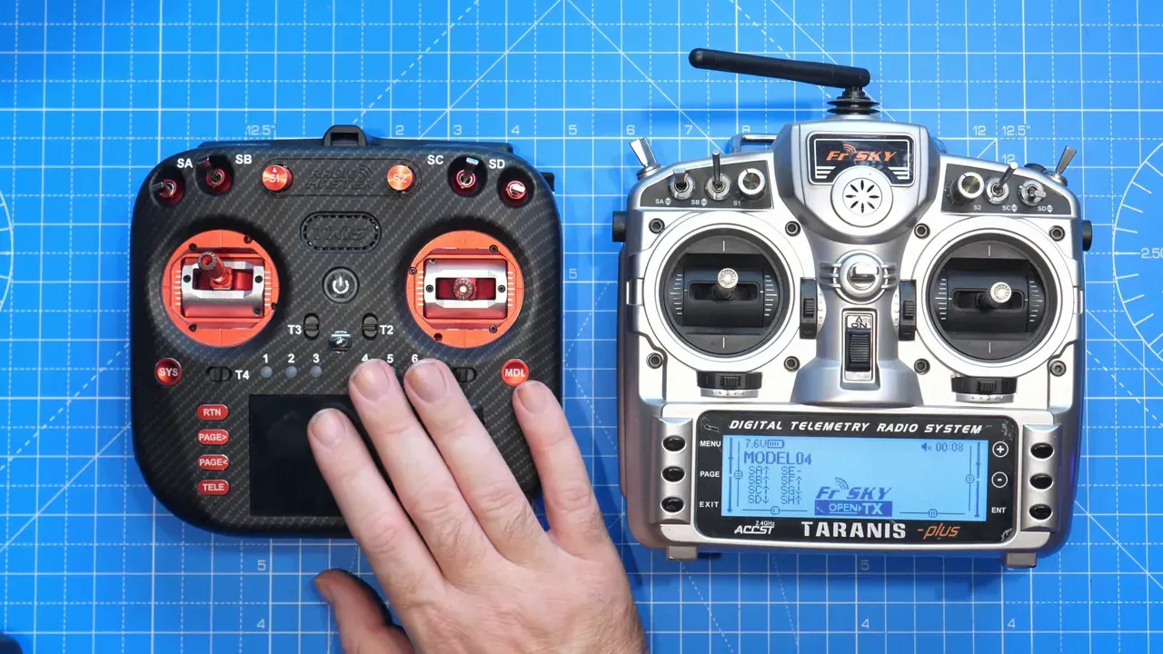 Top-down, high‑clarity image of a carbon‑finish mid‑size transmitter (left) beside a silver FrSky Taranis transmitter (right) on a blue cutting mat with a hand resting on the left unit.
