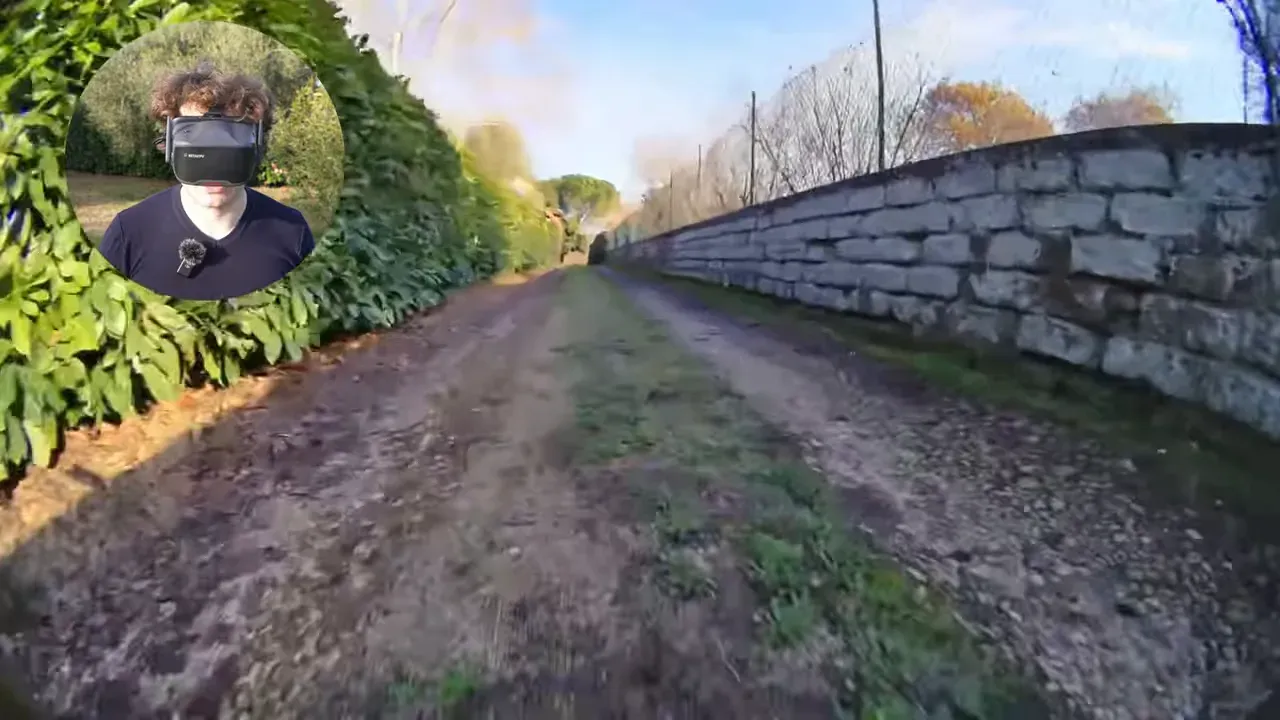 FPV forward view down a dirt track between a tall hedge and a stone wall with presenter inset wearing VR-style goggles