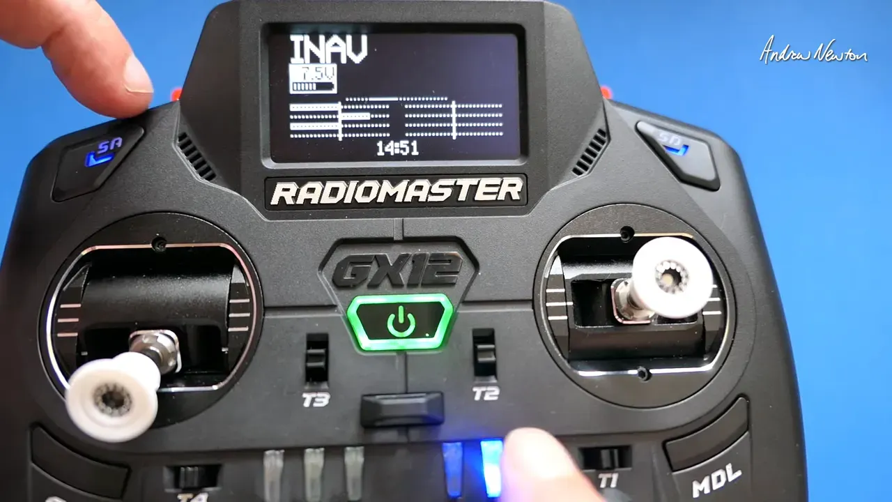 Clear front view of a Radiomaster GX12 with a finger pressing a bottom switch/fader and the LED lit; display, sticks and power button are visible