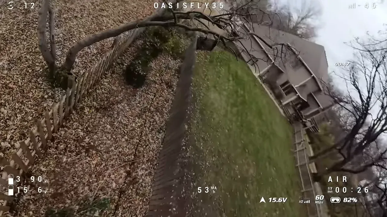 Tilted FPV shot from the OasisFly35 flying low beside a wooden fence and tree with houses in the background and flight telemetry visible