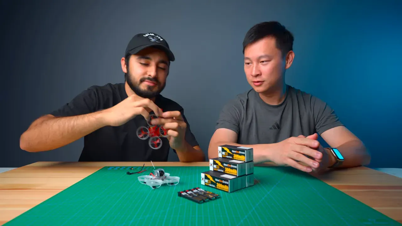 Two presenters at a workbench with stacked Nitro Nectar NX69 300mAh battery boxes, a tiny whoop and small pigtail/connectors on a green cutting mat.