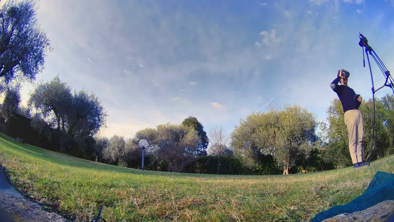 Wide outdoor shot showing presenter wearing FPV goggles and the grassy test field during a hover demonstration