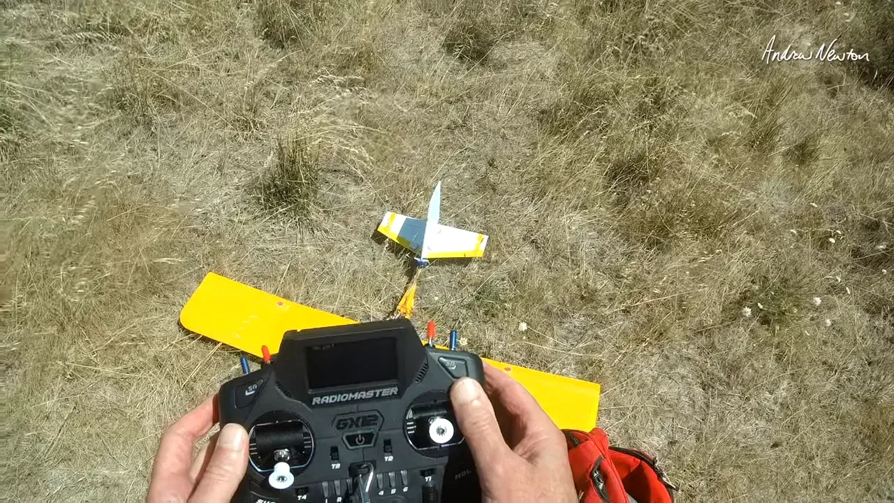 Front view of a Radiomaster GX12 transmitter held over a yellow model airplane on dry grass; the transmitter screen appears dark against the bright background