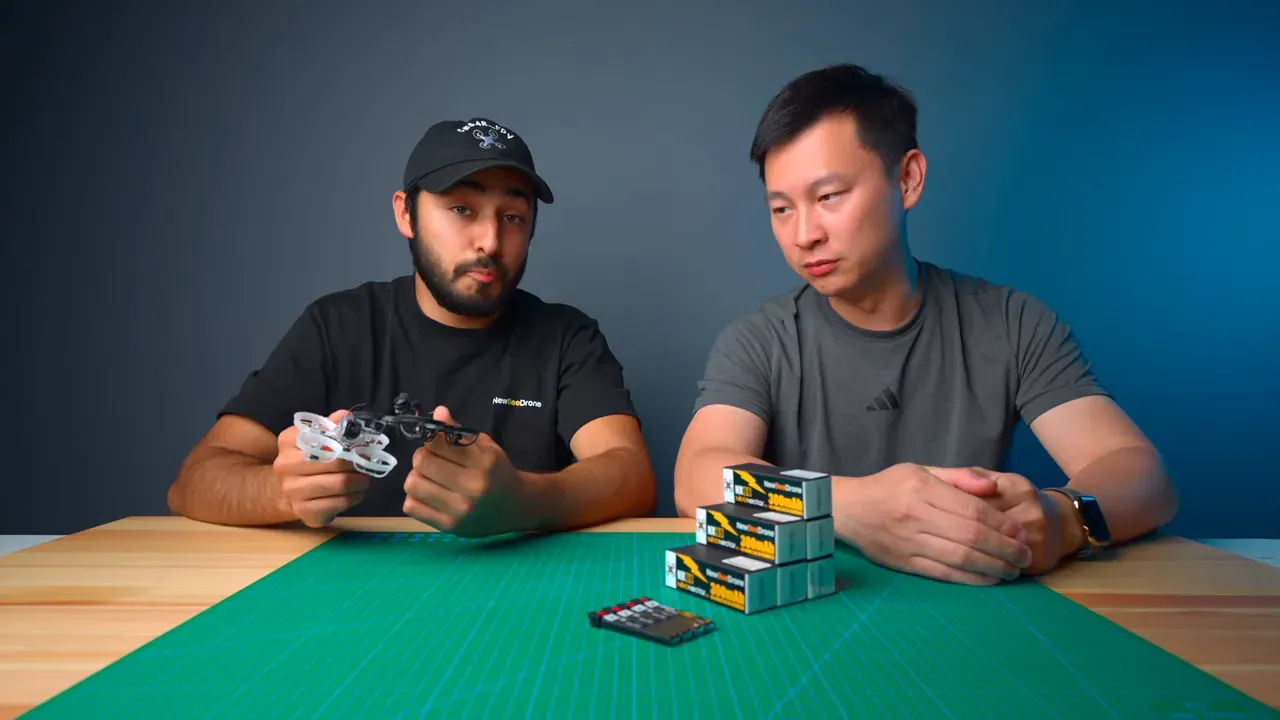 Two presenters hold micro whoops beside a stacked set of Nitro Nectar NX69 300mAh battery boxes on a workbench.