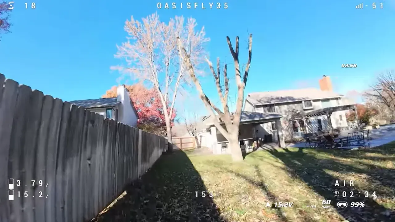 OasisFly35 FPV wide-angle shot of a backyard with a wooden fence, trees and a house under a clear blue sky with flight telemetry overlay