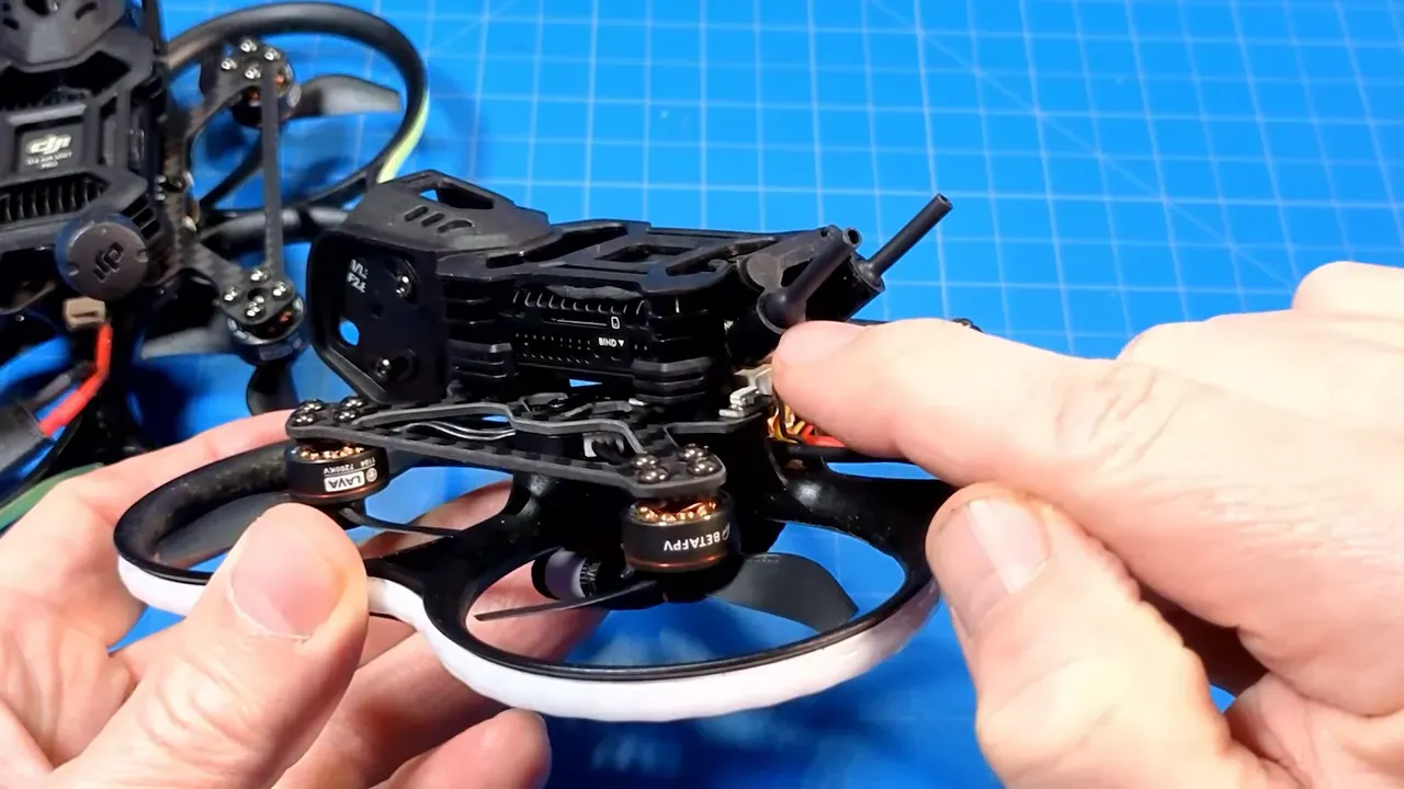Hands holding a Pavo20 Pro II micro quad with a finger indicating the daughterboard/wiring and antenna stubs on the rear of the stack, blue grid work mat background