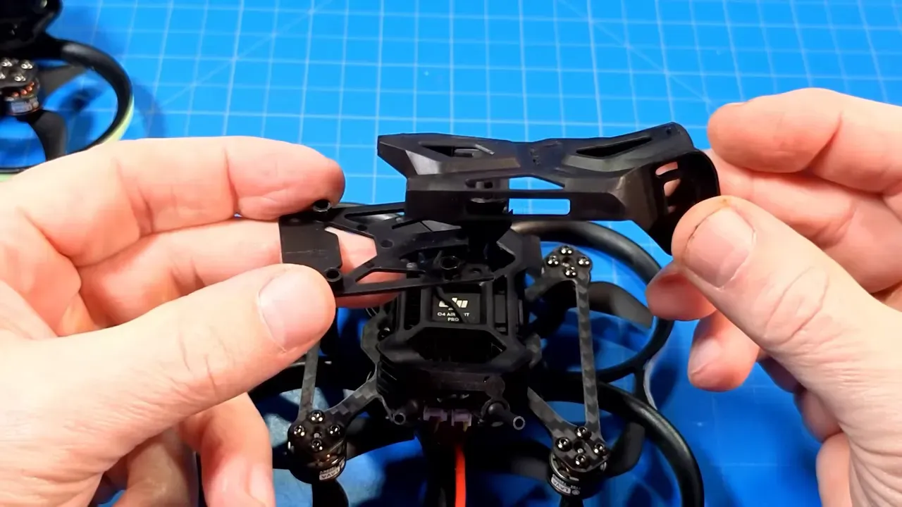 Hands holding two plastic camera mounting brackets above a Pavo20 Pro II frame on a blue work mat, showing how the parts fit together