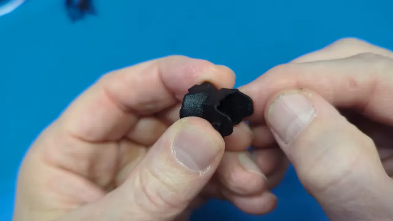 Close-up of hands holding a black 3D‑printed HappyModel FOV extender prototype showing the central aperture, rear latch and printed texture against a blue mat