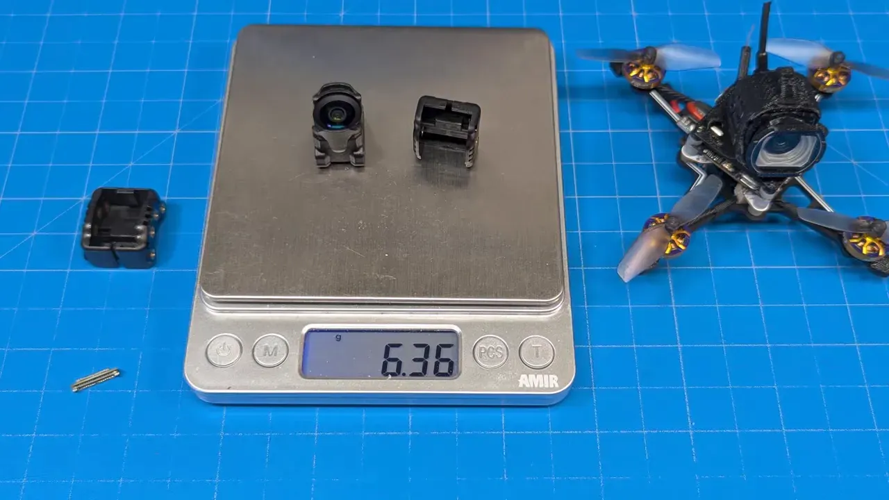 Digital scale showing the FlyWoo wide camera module and its rear housing on the weighing plate with the LCD reading 6.36 grams; a small drone is visible at the right on a blue work mat