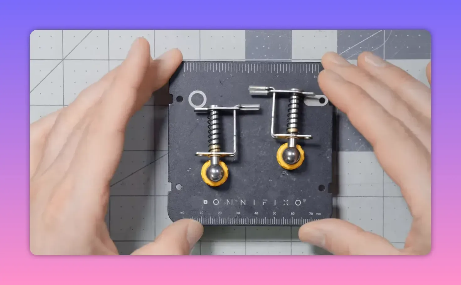 Top-down close-up of an Omnifixo two-clip base with spring-loaded clips and ruler markings, hands framing the unit.