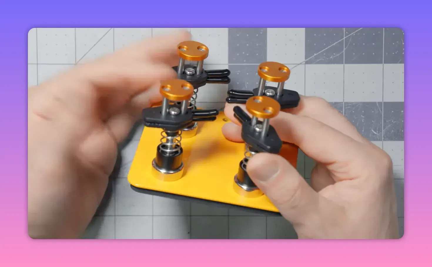 Hands adjusting a compact yellow magnetic helping‑hands base with four spring‑loaded clips on a gridded cutting mat.
