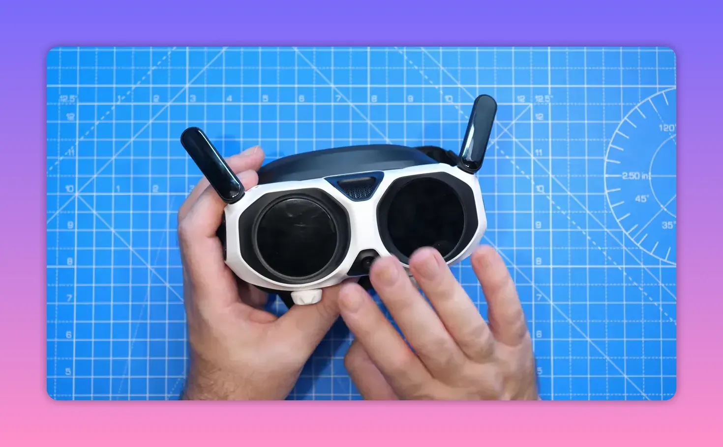 Top-down shot of Antigravity A1 Vision goggles held over a blue grid mat, showing antennas and front housing with reviewer’s hands