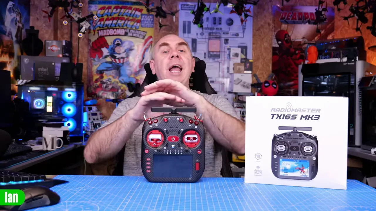 Presenter behind RadioMaster TX16S MK3 radio and boxed packaging on a blue cutting mat, showing front of radio, gimbals and screen