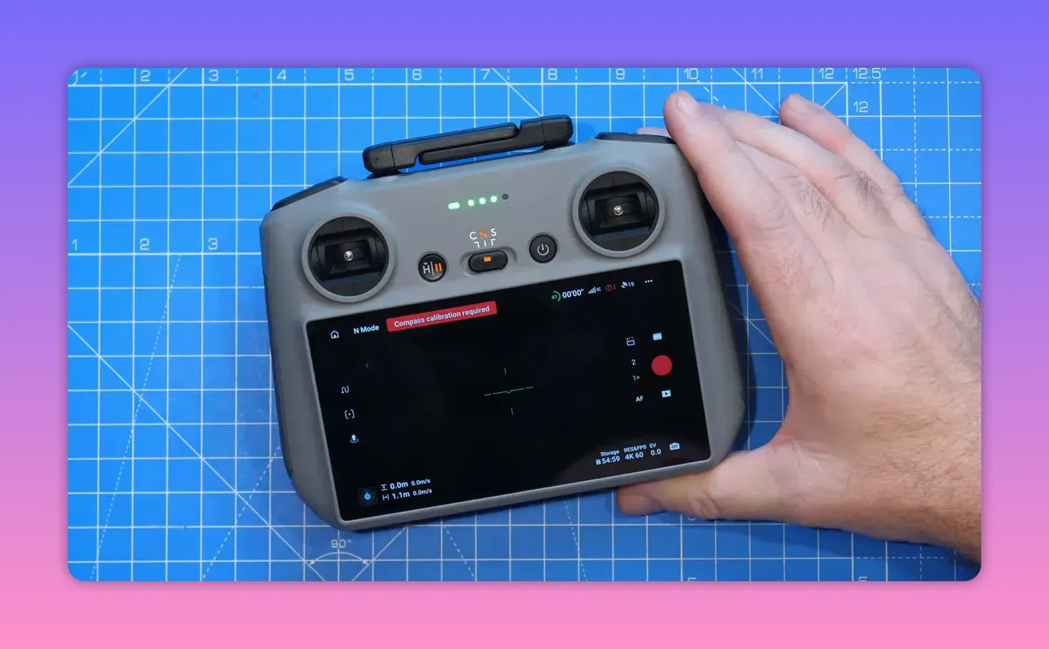 DJI controller on a blue cutting mat with the Fly app visible (red 'Compass calibration required' banner); right hand rests on the edge and the screen is unobstructed.