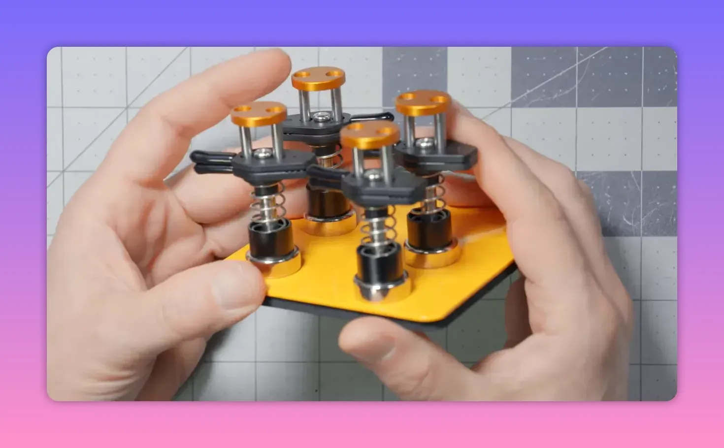 Hands holding a yellow magnetic base with four spring-loaded clips, shown at a slight angle on a gridded cutting mat