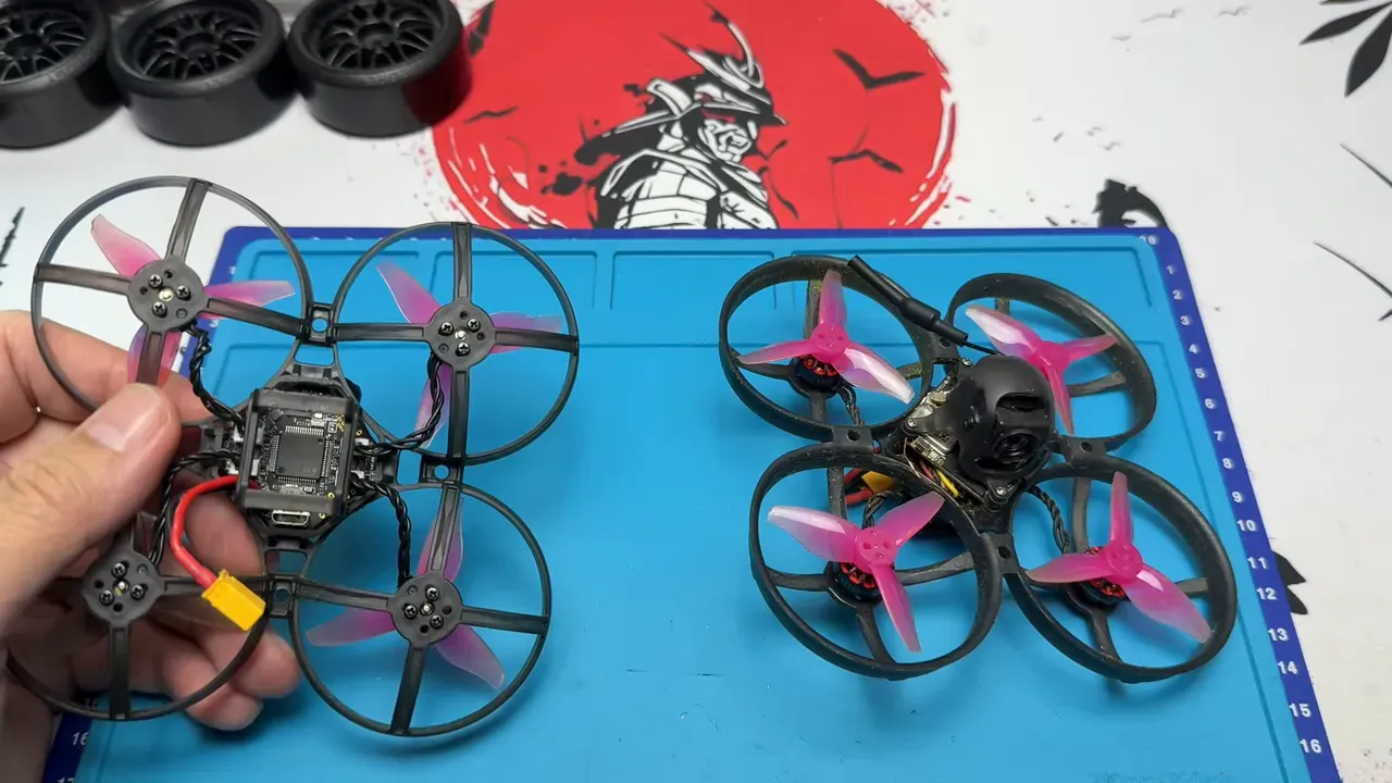 Top-down view of two Mobula-style cinewhoops on a blue work mat, one with canopy and camera visible and the other showing the frame internals