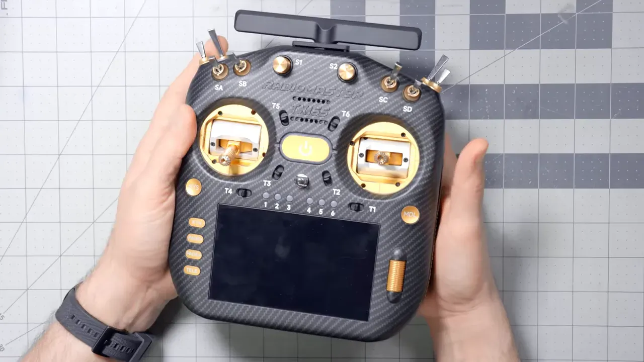 Top-down view of a full-size Radiomaster TX16S-style transmitter with carbon-fiber finish, gold gimbals, multiple switches and a touchscreen held in hands
